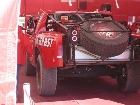 1990 Ford Bronco - Rear bed of a red Terrible Herbst off-road race truck showing a mounted spare tire, taillights and tube bumper in a service area.