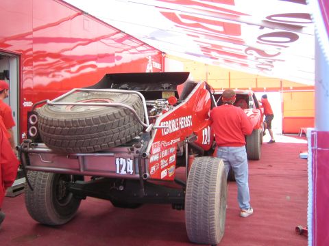 1990 Ford Bronco - Rear of a red number 124 Terrible Herbst off-road racer with twin spare tires under an awning as crew in red shirts work on it.