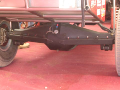 1990 Ford Bronco - Close-up of a heavy black rear axle housing and differential mounted under an off-road race truck on a red carpeted service floor.