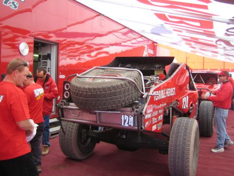 1990 Ford Bronco - Rear view of a red number 124 Terrible Herbst off-road racer with twin mounted spares as crew members stand around it under a team awning.