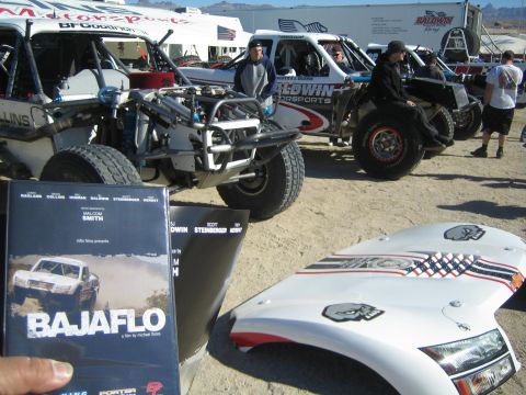 1990 Ford Bronco - hand holds a BAJAFLO film DVD in front of a row of off-road race buggies and a white body panel at a desert race paddock.