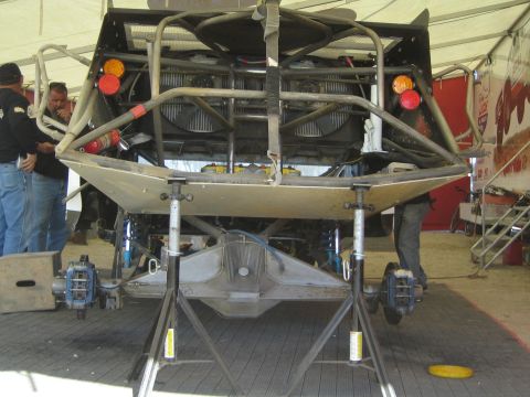 1990 Ford Bronco - Rear of an off-road race vehicle raised on jack stands under an awning, exposing the bare tube chassis, radiator and brake assemblies.