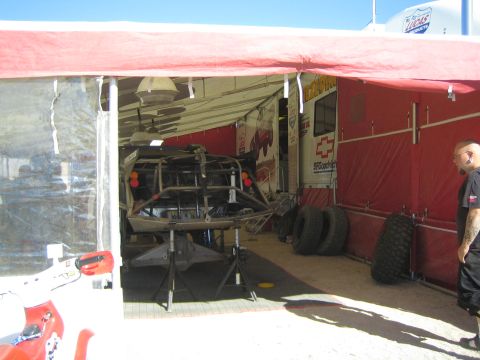 1990 Ford Bronco - off-road race chassis raised on jack stands inside a red Lucas Oil team service tent with spare tires stacked along the wall.
