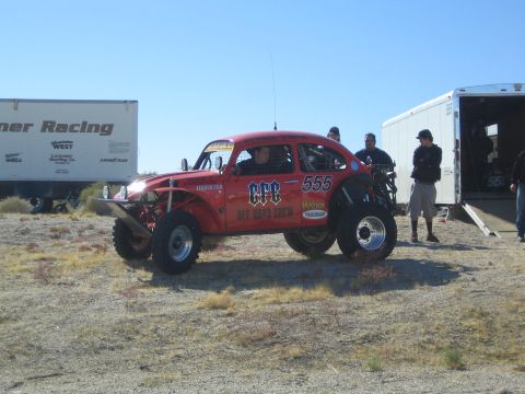1990 Ford Bronco - Red CPC Off Road Crew number 555 Baja Bug off-road racer on tall tires parked in the desert beside team trailers with crew nearby.