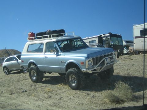 1990 Ford Bronco - Light blue early-1970s Chevrolet Blazer with a roof rack and roll bar, lifted on off-road tires, parked in a desert lot beside an RV.