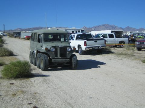 1990 Ford Bronco - Olive-drab six-wheeled Willys-based military-style vehicle driving on a desert dirt road past parked trucks and trailers.