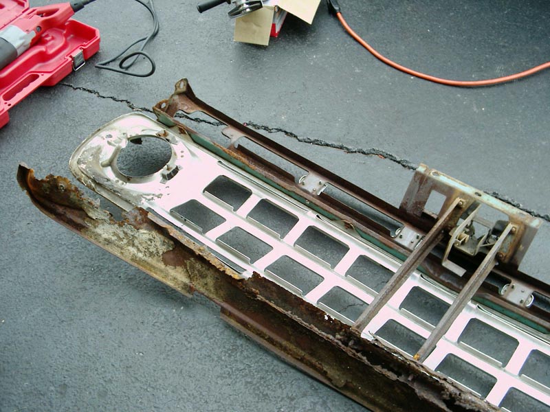 Rear of white truck grille with empty headlight hole, hood latch and rusty support on asphalt