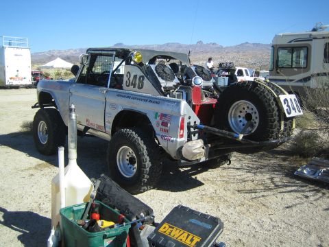 1990 Ford Bronco - silver early Ford Bronco race truck marked #348 sits in a desert staging area beside RVs, with tools and a spare tire nearby.