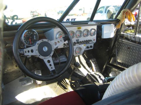 1990 Ford Bronco - stripped interior of a race vehicle shows a bank of aftermarket gauges, a dished steering wheel, floor shifter and roll cage tubing.