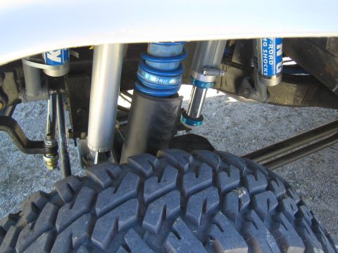 1990 Ford Bronco - detailed view of an off-road truck's front suspension showing dual blue bypass and coilover shocks above a knobby tire.