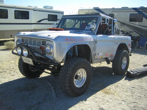 1990 Ford Bronco - lifted silver early Ford Bronco race truck #348 with West Coast Broncos lettering sits among RVs in a desert pit area.