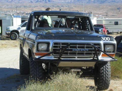1990 Ford Bronco - Front view of a black square-body Ford off-road race truck #300 with a tube bumper and occupants inside, mountains behind.