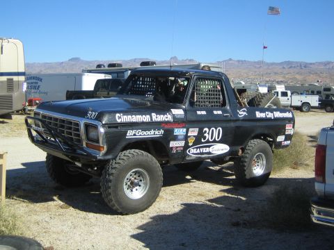 1990 Ford Bronco - black square-body Ford race truck #300 covered in sponsor decals including BFGoodrich and Cinnamon Ranch parked in a desert lot.