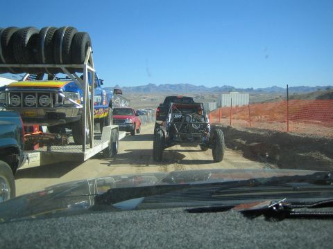 1990 Ford Bronco - Shot through a windshield, off-road vehicles and a trailer stacked with tires travel a dirt road in a desert landscape.