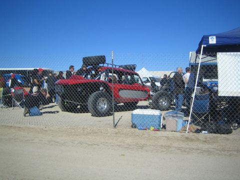 1990 Ford Bronco - red open-cockpit off-road race buggy sits behind a chain-link fence as people and a canopy tent gather in the pit area.
