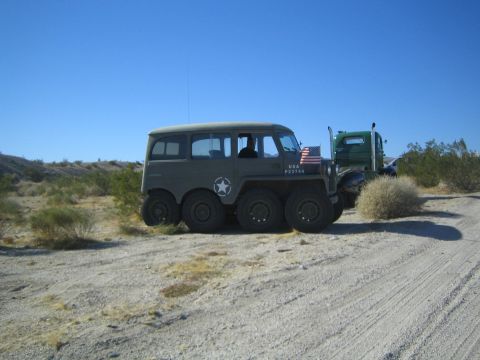 1990 Ford Bronco - unusual olive-drab military-style vehicle riding on eight wheels and marked USA sits on a desert dirt road near a green truck.