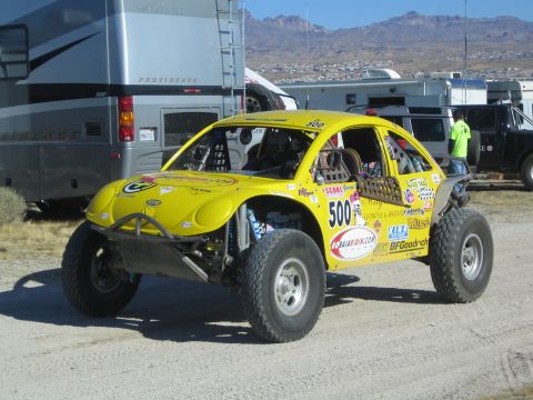 1990 Ford Bronco - yellow VW-style Baja Bug off-road race car #500 covered in sponsor decals stands on a dirt lot beside motorhomes.
