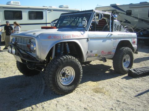 1990 Ford Bronco - lifted silver early Ford Bronco race truck #348 lettered West Coast Broncos sits among RVs with onlookers in a desert lot.
