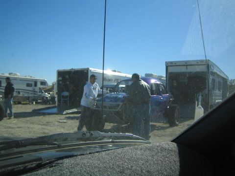 1990 Ford Bronco - Seen through a dusty windshield, people work around an off-road buggy parked between two enclosed trailers in a desert lot.