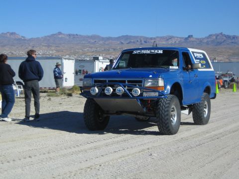 1990 Ford Bronco - blue lifted Ford Bronco off-road truck with a tube bumper and roof light bar idles on a desert pit road as people watch.