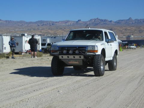 1990 Ford Bronco - white lifted Ford Bronco off-road truck with a tube front bumper drives a desert dirt road past trailers and spectators.