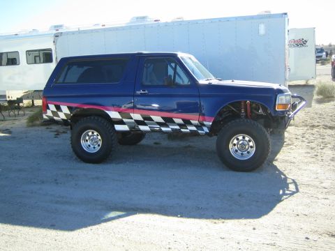 1990 Ford Bronco - blue full-size Ford Bronco with black-and-white checkered side graphics and a pink stripe stands lifted on a dirt lot by trailers.