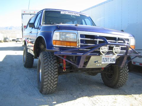 1990 Ford Bronco - Front view of a blue lifted Ford Bronco with a tube bumper, Hella driving lights and a California DZRT RUN plate in the desert.