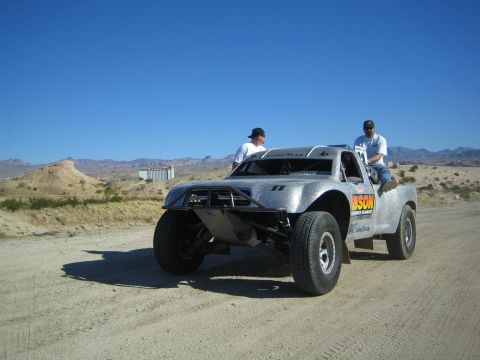 1990 Ford Bronco - silver tube-frame Ford prerunner trophy truck sits on a desert dirt road as two men climb aboard under blue sky.