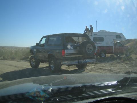1990 Ford Bronco - black Ford Bronco with a rear-mounted spare tire tows a white camper trailer across the desert as two men ride atop it.