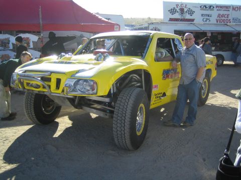 1990 Ford Bronco - man stands beside a bright yellow Ford tube-frame trophy truck parked at a desert race pit lined with vendor banners.
