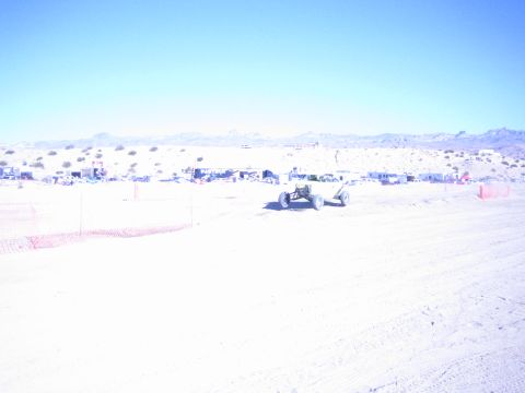 1990 Ford Bronco - single offroad buggy crosses a vast washed-out desert race course with distant pit camps and mountains beyond.