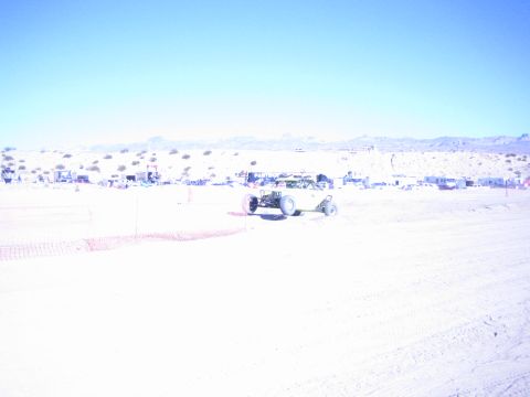 1990 Ford Bronco - offroad race car runs across a sun-washed desert course with a row of pit camps and mountains on the horizon.