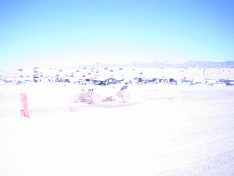 1990 Ford Bronco - offroad race car runs behind orange safety fencing on a sun-bleached desert course with pit camps in the distance.