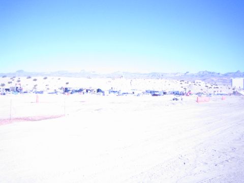 1990 Ford Bronco - Distant pit camps and vehicles spread across a wide sun-bleached desert race course below a rocky mountain range.