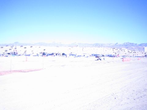 1990 Ford Bronco - wide washed-out desert race course stretches toward distant pit camps and orange fencing beneath low desert hills.
