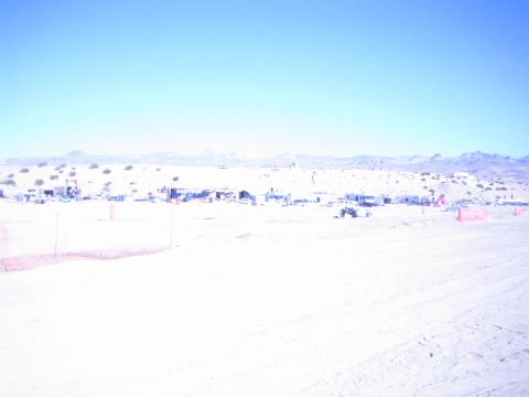 1990 Ford Bronco - sun-bleached desert race area shows scattered pit camps and orange safety fencing below a distant mountain range.