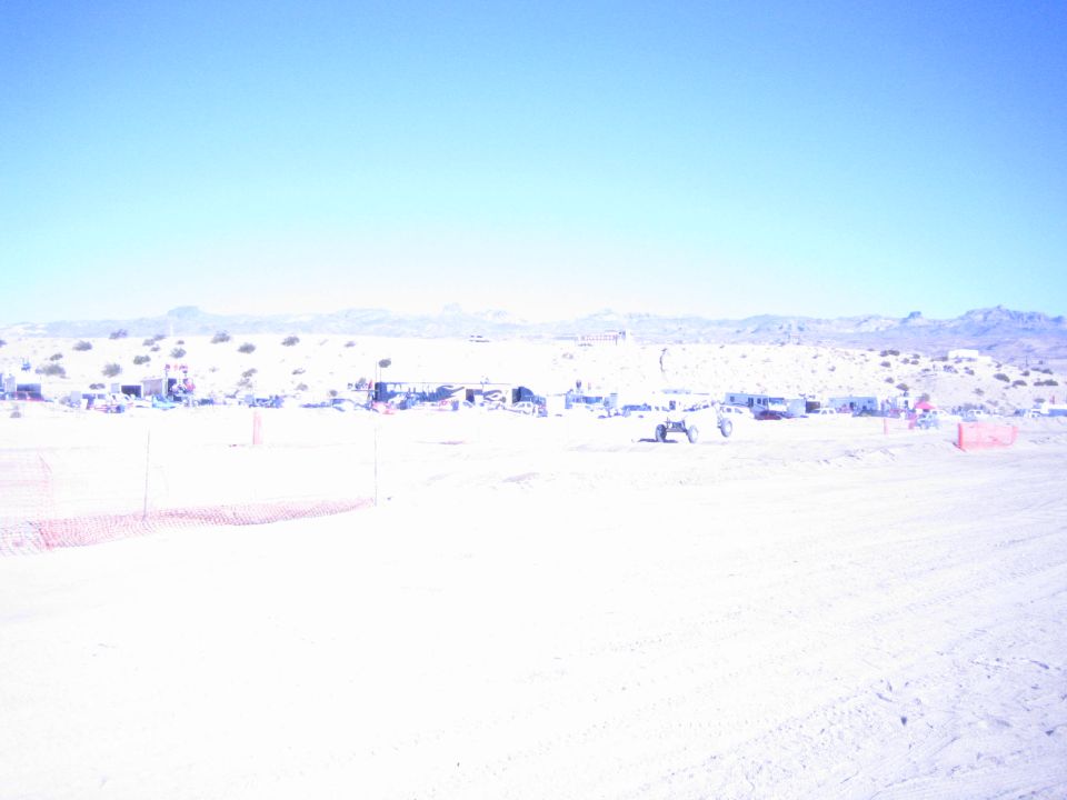 Overexposed desert race course with distant pit camps and orange fencing