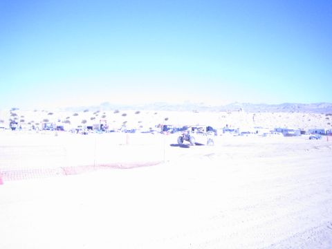 1990 Ford Bronco - offroad buggy crosses a sun-bleached desert race course past scattered pit camps below a distant mountain range.