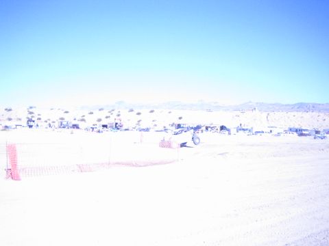 1990 Ford Bronco - distant offroad vehicle and a row of pit camps sit along a sun-bleached desert course lined with orange fencing.