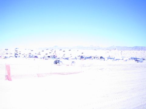 1990 Ford Bronco - offroad vehicle runs behind orange safety fencing across a wide sun-bleached desert course with distant pit camps.