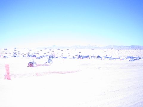 1990 Ford Bronco - offroad buggy kicks up dust behind orange fencing on a sun-bleached desert course lined with distant pit camps.