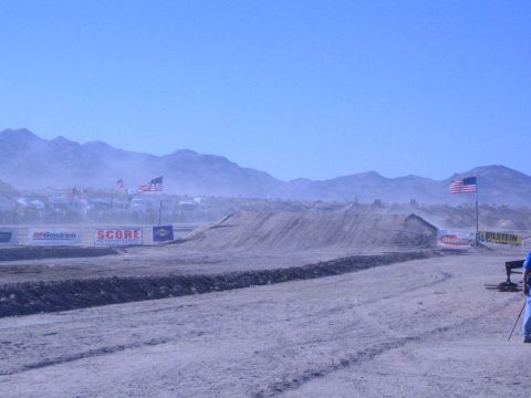1990 Ford Bronco - dusty desert race course features a graded dirt berm, American flags and sponsor banners below a mountain range.