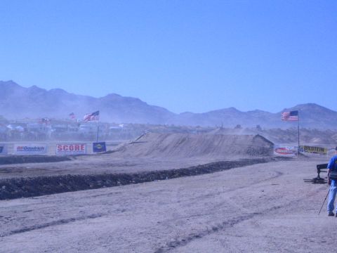 1990 Ford Bronco - man watches a dusty desert race course lined with sponsor banners and American flags below distant mountains.