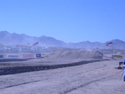 1990 Ford Bronco - dusty desert race course shows a graded dirt berm, American flags and sponsor banners beneath a mountain range.