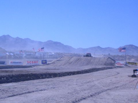 1990 Ford Bronco - Offroad vehicles crest a dirt berm on a dusty desert race course lined with sponsor banners and American flags.
