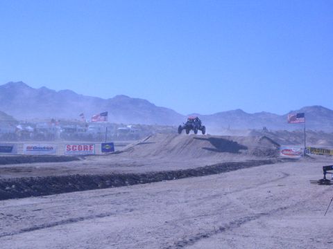 1990 Ford Bronco - open-wheel off-road race buggy launches off a dirt jump at a desert course lined with American flags and SCORE and BFGoodrich banners.