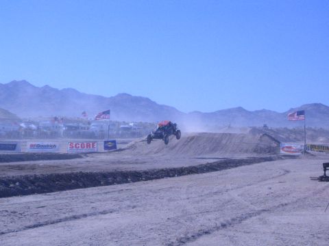 1990 Ford Bronco - off-road race buggy is airborne over a dirt jump on a dusty desert course backed by mountains, flags, and sponsor banners.