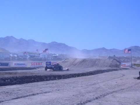 1990 Ford Bronco - off-road race buggy lands on a dirt jump at a dusty desert course with American flags, mountains, and SCORE banners behind.