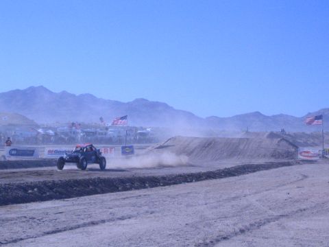 1990 Ford Bronco - red and white off-road race buggy throws up a cloud of dust as it races along a desert dirt course lined with flags and banners.