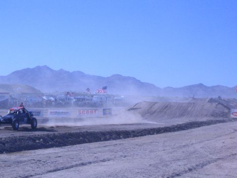 1990 Ford Bronco - off-road race buggy speeds along a desert course past a dirt berm, with spectators, flags, and banners in a dusty background.